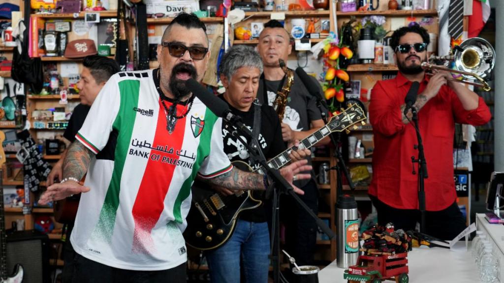 Un grupo de músicos en un escenario pequeño, interpretando música. Uno de ellos lleva una camiseta con los colores de la bandera palestina, mientras toca un micrófono. Hay un guitarrista, un saxofonista y un trompetista, rodeados de varios objetos decorativos en el fondo.