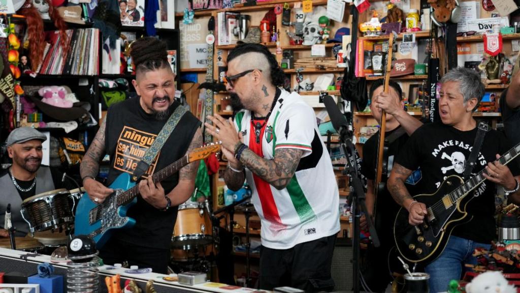 Un grupo de músicos en un escenario pequeño, tocando instrumentos como guitarra y batería, mientras interactúan y se divierten entre ellos. El fondo está decorado con diversos objetos coloridos.