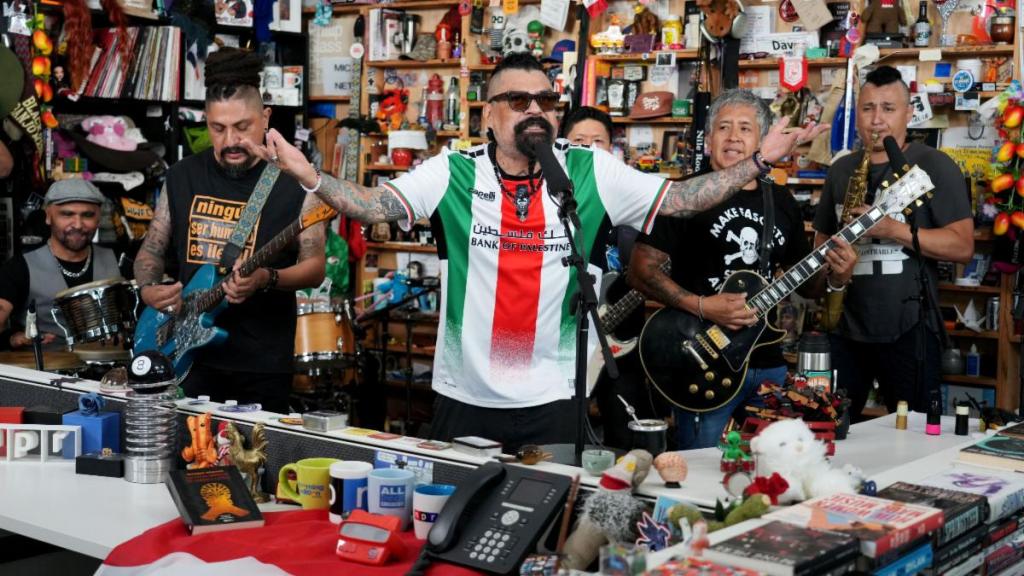 Grupo musical en una actuación en vivo en un entorno lleno de objetos coloridos y decoraciones, con instrumentos como guitarras, batería y saxofón, mientras el vocalista gesticula animadamente.